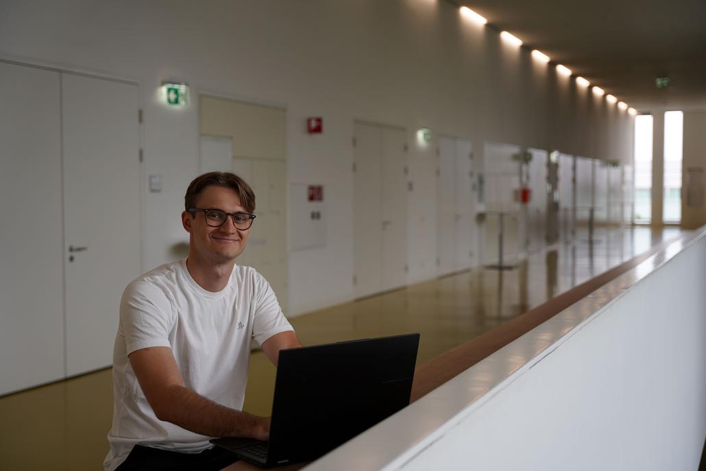 Mann mit Brille und weißem T-Shirt arbeitet am Laptop in einem modernen Gang mit vielen Türen.